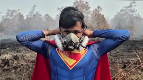 Teenage boy wearing a superman costume and gas mask standing in land burnt by wildfires